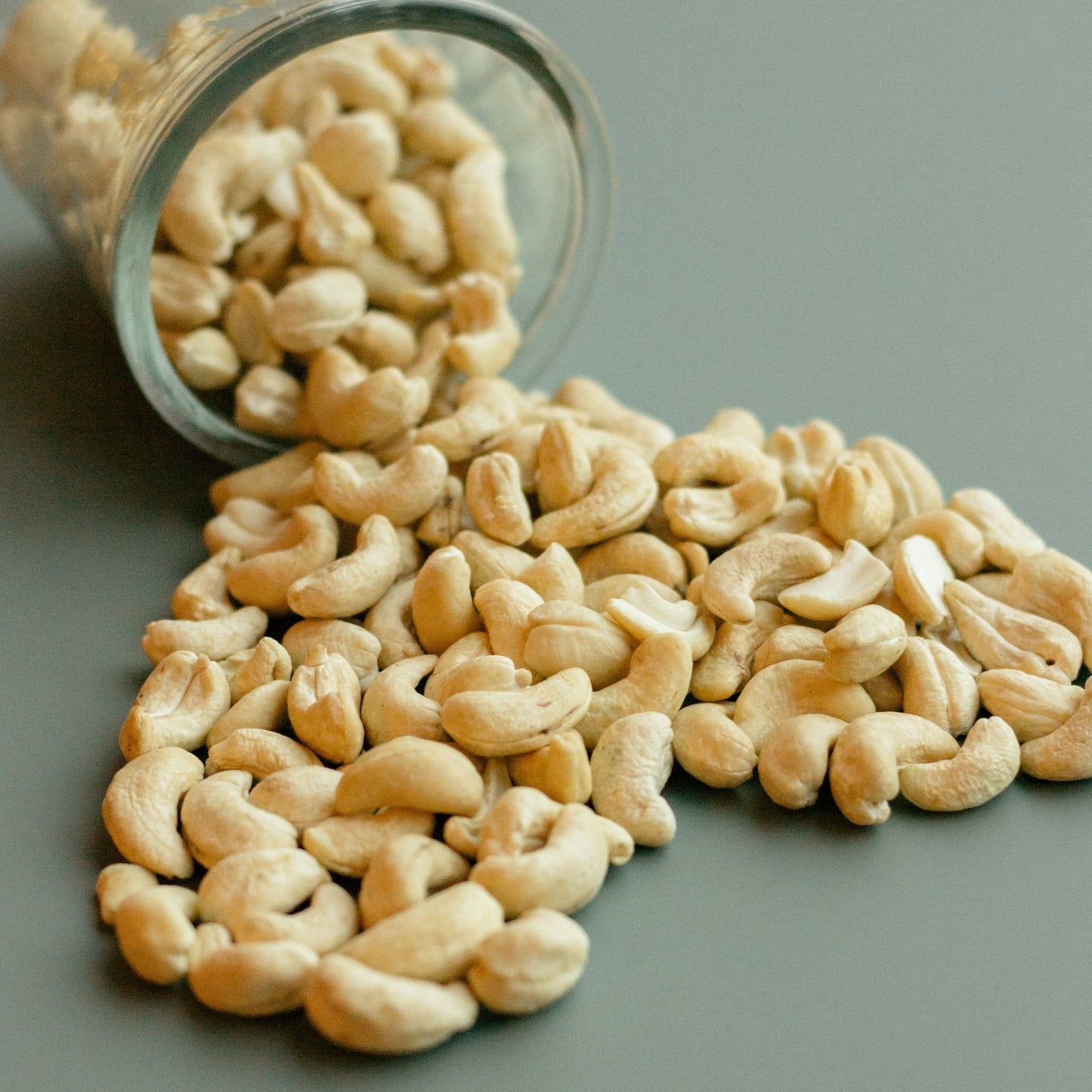 Et væltet glas fyldt med cashewnødder samt en masse cashewnødder spredt ud over på et bord.